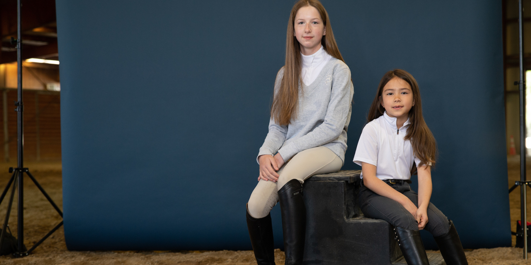 Two young girls in riding clothes sitting on a mounting block. Photographed with a navy background in a riding arena.