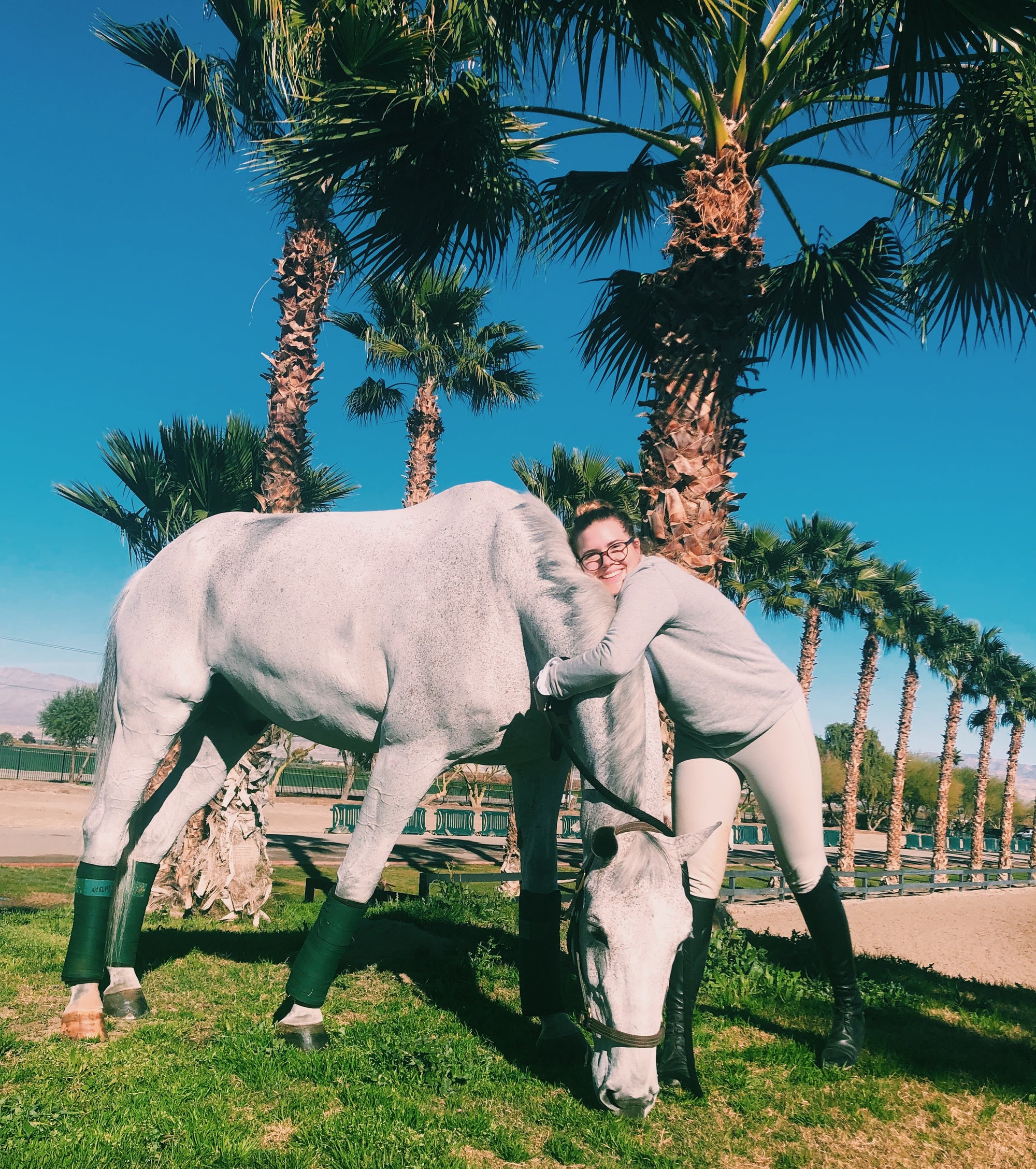 Girl lets grey horse graze while she hugs his neck and smiles at the camera. She's dressed in a grey sweatshirt, tan breeches and Parlanti tall boots.