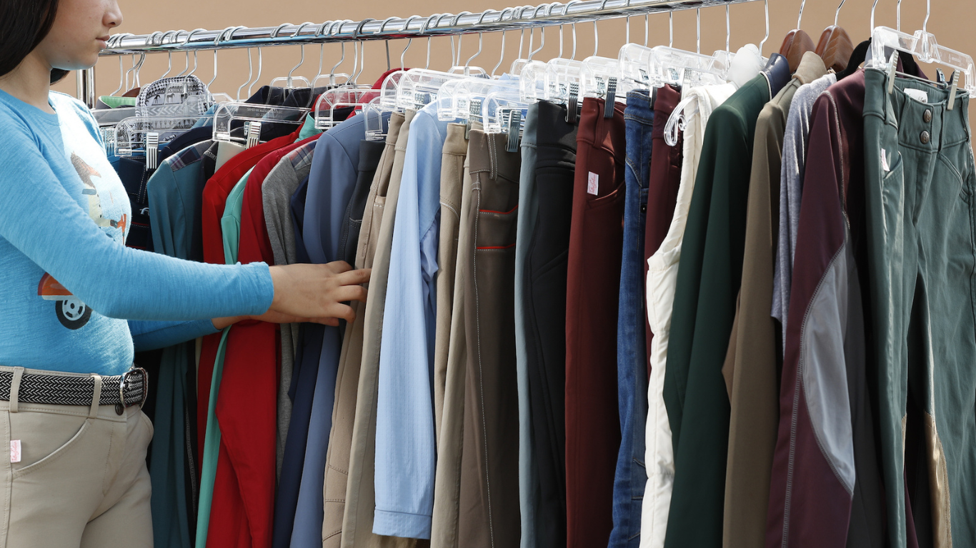 Girl looks through rack of equestrian apparel including Tailored Sportsman breeches, sunshirts, and Equiline show jackets.