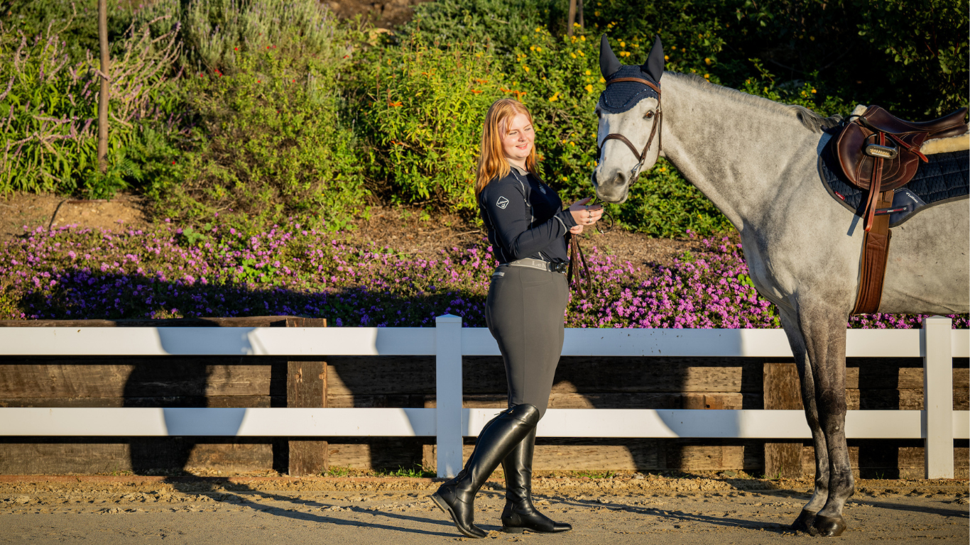Girl hold grey horse wearing a Lemieux base layer and grey breeches and tall boots. Horse has matchy navy set with CWD saddle.