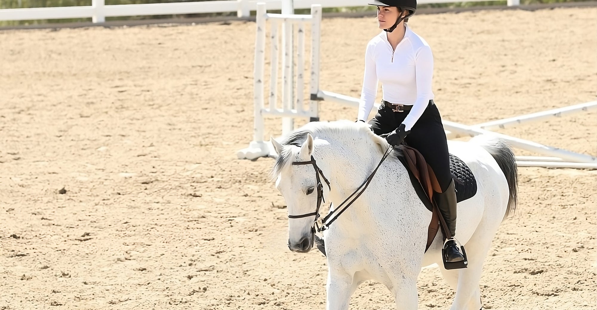 Girl in white sunshirt and black breeches trots on a grey horse in an arena.